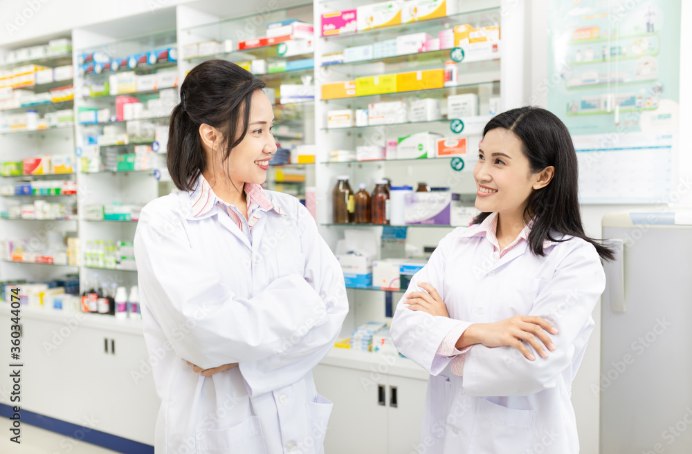 two Asian healthcare stand and cross arms in drugstore, unity and teamwork, happiness organization, asian female pharmacist feeling happy in work time