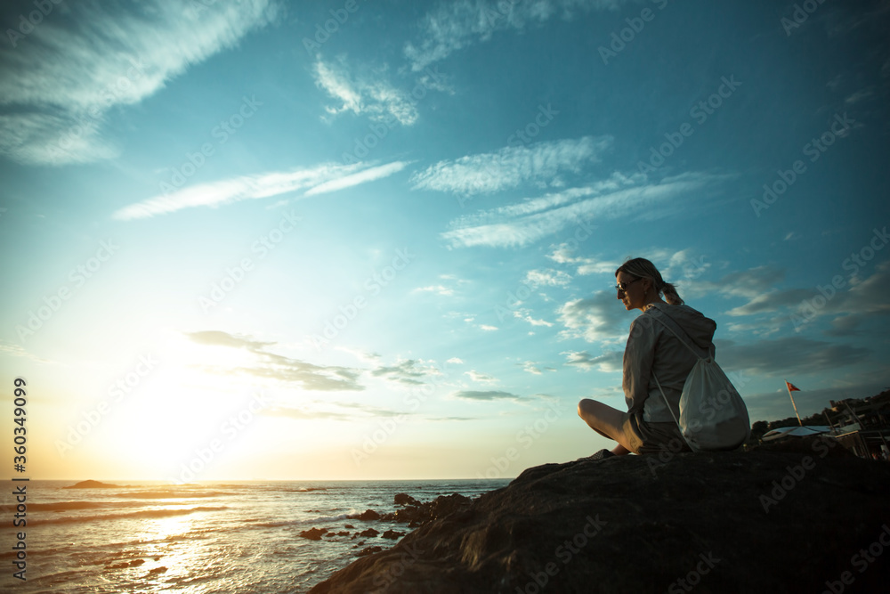 Young woman sitting on the rocky ocean coast during sunset.