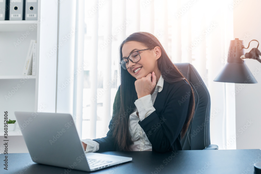 Young happy businesswoman sitting in her bright office while looking at the laptop. Young happy businesswoman sitting in her bright office while looking at the laptop.
