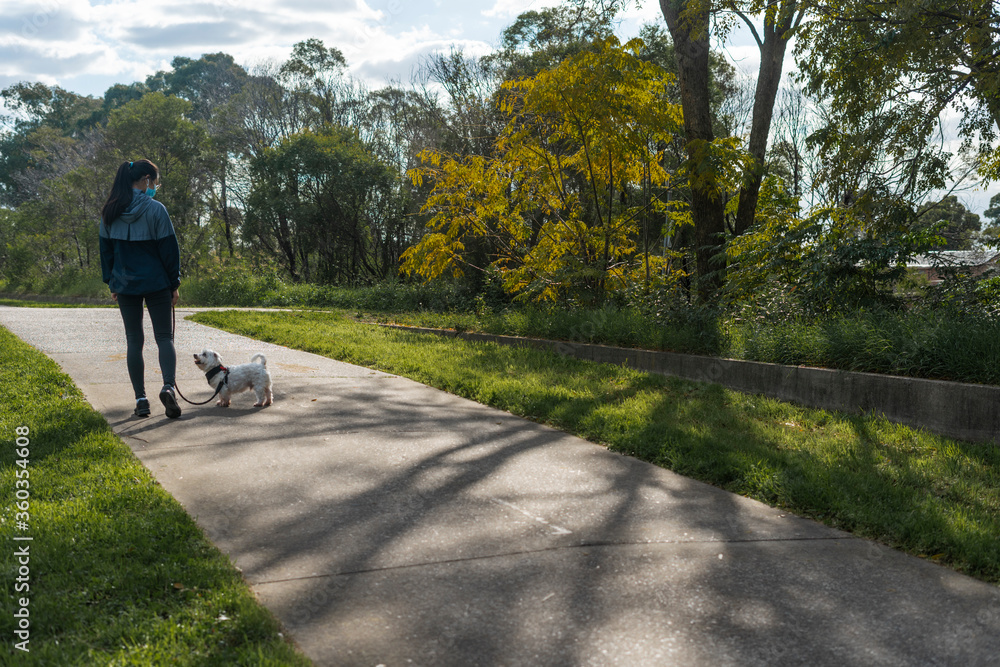 Girl in surgical mask in casual urban athletic wear walking with pet dog on a leash at a nature park on a footpath stylized to highlight mask during covid-19 pandemic social distancing isolation Girl in surgical mask in casual urban athletic wear walking with pet dog on a leash at a nature park on a footpath stylized to highlight mask during covid-19 pandemic social distancing isolation