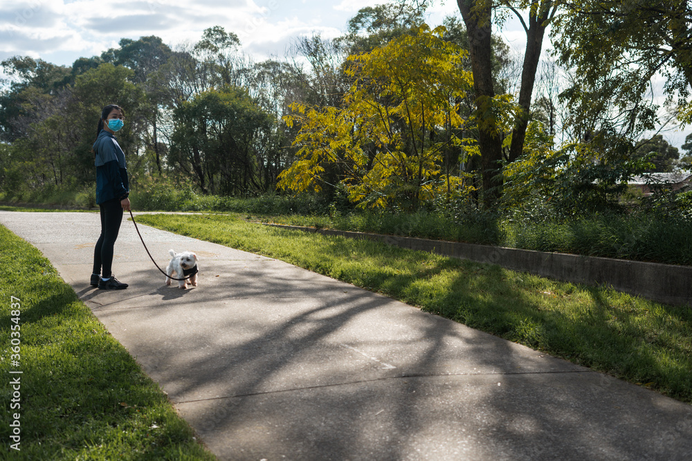 Obraz premium Girl in surgical mask in casual urban athletic wear walking with pet dog on a leash at a nature park on a footpath stylized to highlight mask during covid-19 pandemic social distancing isolation