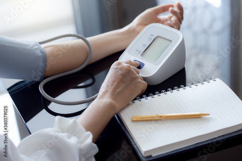 Obraz na plátně Woman measuring blood pressure by using digital sphygmomanometer with empty white notebook or diary on the desk at home