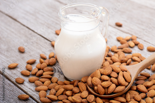 Almond milk in glass pitcher and almond nuts in wooden spoon on wood table background.