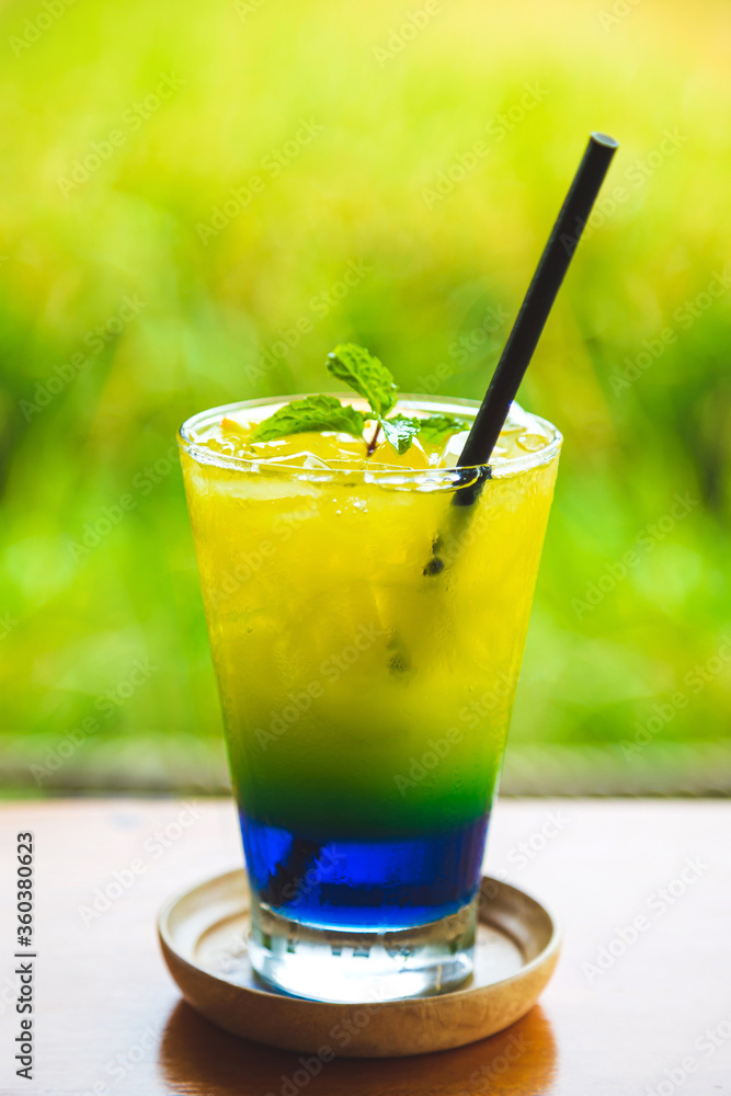 Vertical image a cup of yellow and blue beverage with peppermint on a wooden table with a light green nature blurred background. Vertical image a cup of yellow and blue beverage with peppermint on a wooden table with a light green nature blurred background.