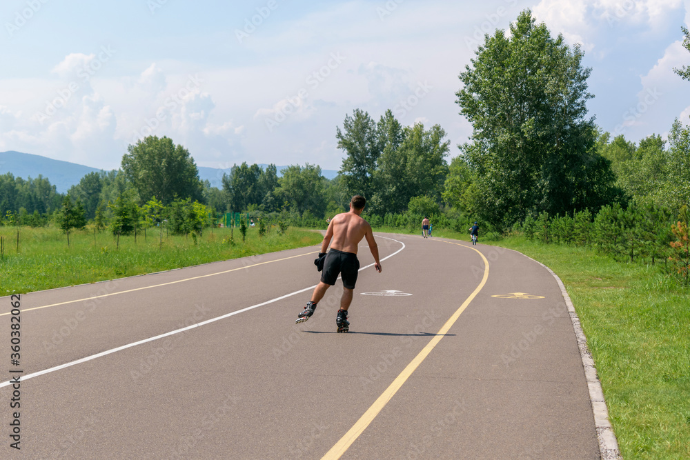 A man in shorts is rollerblading in a park. Asphalt path, green trees and grass. View from the back, muscular physique. Concept of a healthy lifestyle, sports, summer vacations.