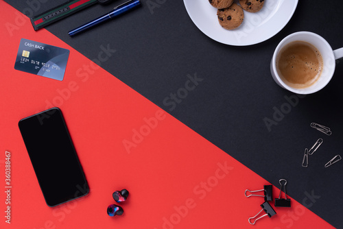 Top view - Office desk workplace with coffee cup, cookies, smartphone, and credit card on red and black background.