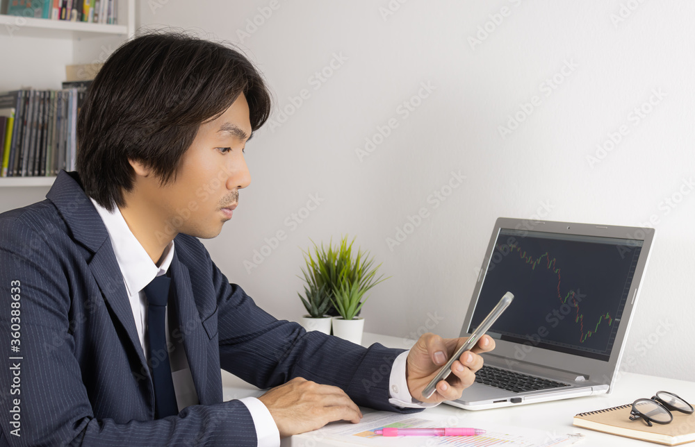 Young Asian Forex Trader or Investor or Businessman in Suit Looking Smartphone and Trading Forex or Stock Chart by Laptop in Trader Room Young Asian Forex Trader or Investor or Businessman in Suit Looking Smartphone and Trading Forex or Stock Chart by Laptop in Trader Room