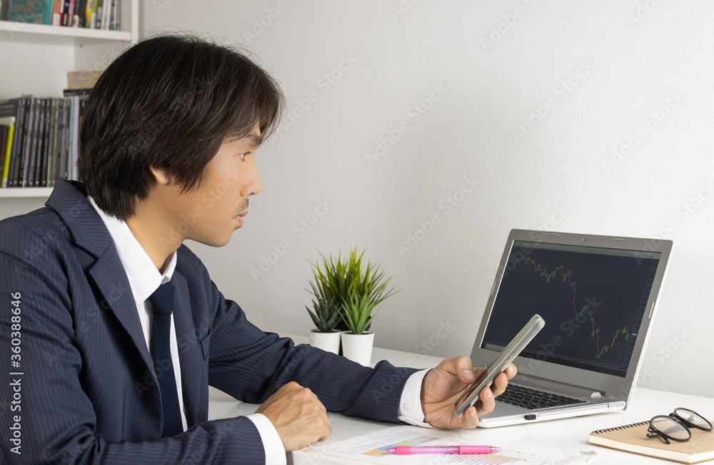 Young Asian Forex Trader or Investor or Businessman in Suit Hold Smartphone and Trading Forex or Stock Chart by Laptop in Trader Room Young Asian Forex Trader or Investor or Businessman in Suit Hold Smartphone and Trading Forex or Stock Chart by Laptop in Trader Room