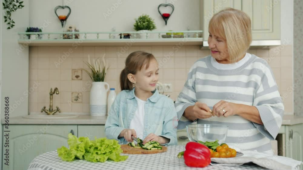 Cute little girl and elderly woman granny are cooking salad at kitchen table together preparing meal and talking. Lifestyle, nutrition and people concept. Cute little girl and elderly woman granny are cooking salad at kitchen table together preparing meal and talking. Lifestyle, nutrition and people concept.
