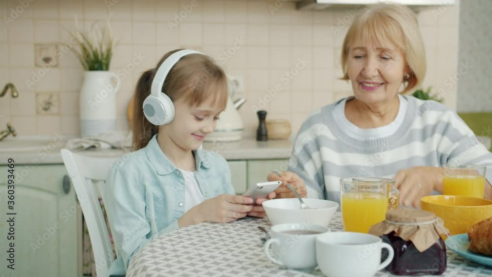 Slow motion of happy little girl using smartphone listening to music in headphones at kitchen table with granny old lady mixing food in bowl Slow motion of happy little girl using smartphone listening to music in headphones at kitchen table with granny old lady mixing food in bowl