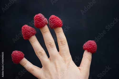 Ripe large raspberries are dressed on the fingers of the hands. Like funny hats. Hand with berries on a black background.
