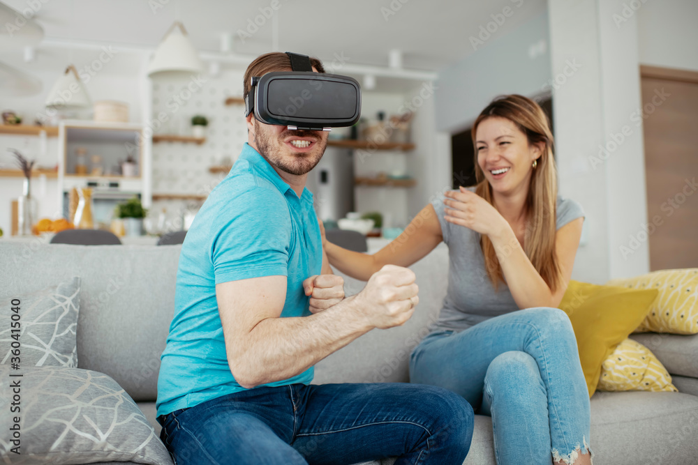 Fototapeta premium Smiling young man using VR headset at home on couch. Man and his wife enjoying virtual reality at her apartment. 