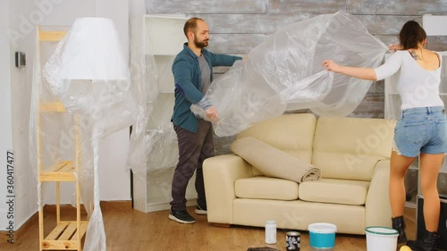 Couple wrapping sofa in plastic foil for protection while they are renovate living room. Home renovation, construction, painting work.