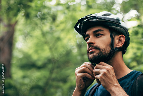Closeup portrait of sporty young man close the protective helmet and looking away, posing on nature background. Male athlete preparing before the riding the bike in the mountain. © iuricazac
