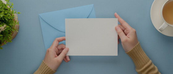 Female hands holding mock-up greeting card above blue envelope on light blue desk