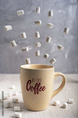 Beige cup of coffee with lettering standing on light wooden table with white flying marshmallows at kitchen