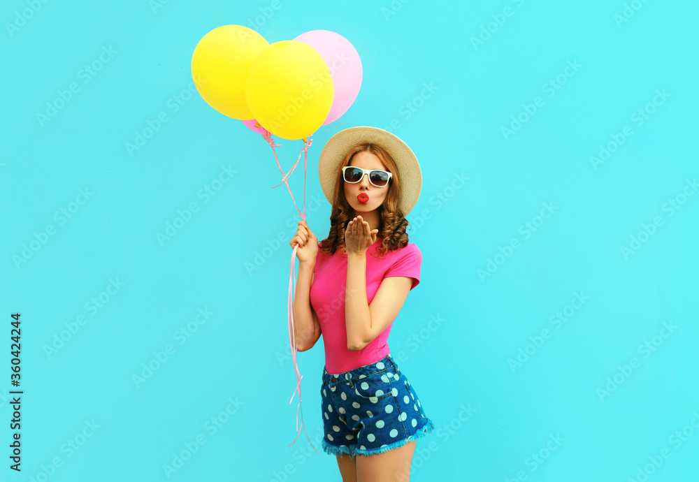 Summer colorful image of cute young woman with yellow pink balloons blowing red lips sending sweet air kiss wearing a shorts and straw hat on blue wall background Summer colorful image of cute young woman with yellow pink balloons blowing red lips sending sweet air kiss wearing a shorts and straw hat on blue wall background