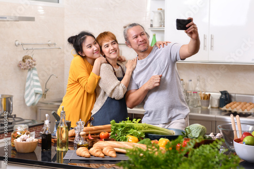 Happy family of mother father and daughter cooking in kitchen making healthy food together and using smart phone to take selfies