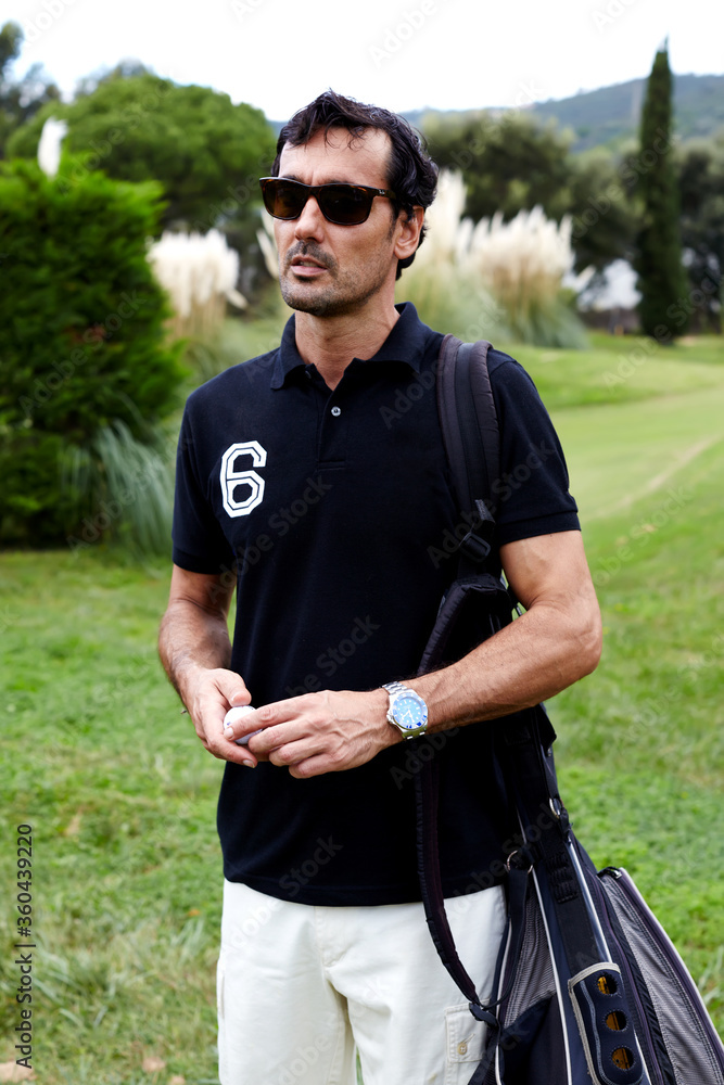 Portrait of stylish golfer man in glasses standing on beautiful golf course looking away, handsome brunette hair man with golf bag on shoulder holding ball in the hands Portrait of stylish golfer man in glasses standing on beautiful golf course looking away, handsome brunette hair man with golf bag on shoulder holding ball in the hands