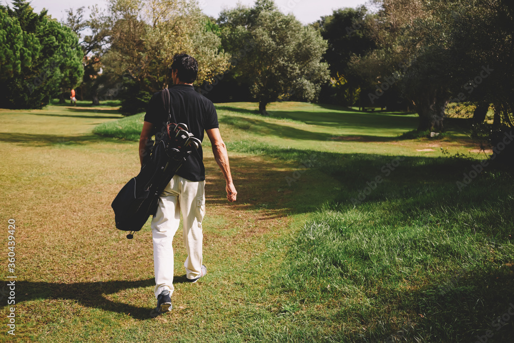 Rear view of professional male golfer walking to the next hole in golf course, good golf game at sunny summer day on the course Rear view of professional male golfer walking to the next hole in golf course, good golf game at sunny summer day on the course