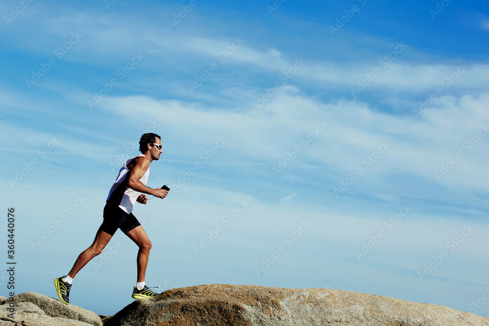 Sporty male runner jogging outdoors against copy space sky background, mature male engaged in running while listening to music in headphones, working out in mountain landscape Sporty male runner jogging outdoors against copy space sky background, mature male engaged in running while listening to music in headphones, working out in mountain landscape