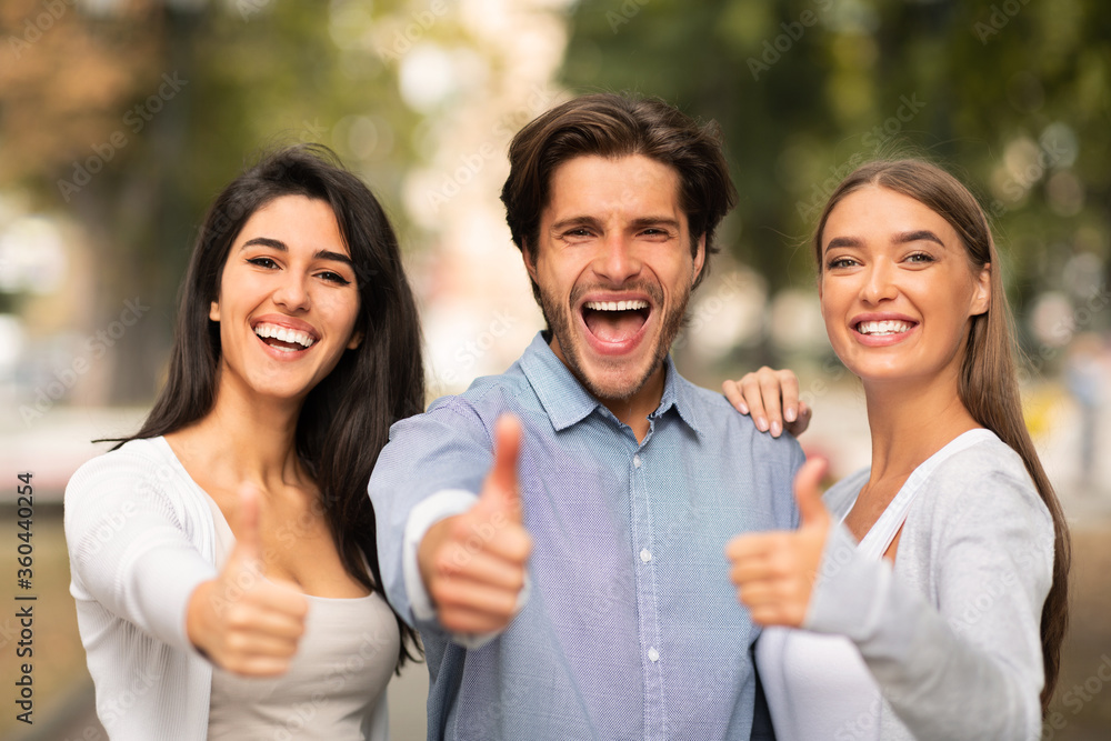 Three Cheerful Friends Gesturing Thumbs-Up Approving Something Standing Outside Three Cheerful Friends Gesturing Thumbs-Up Approving Something Standing Outside