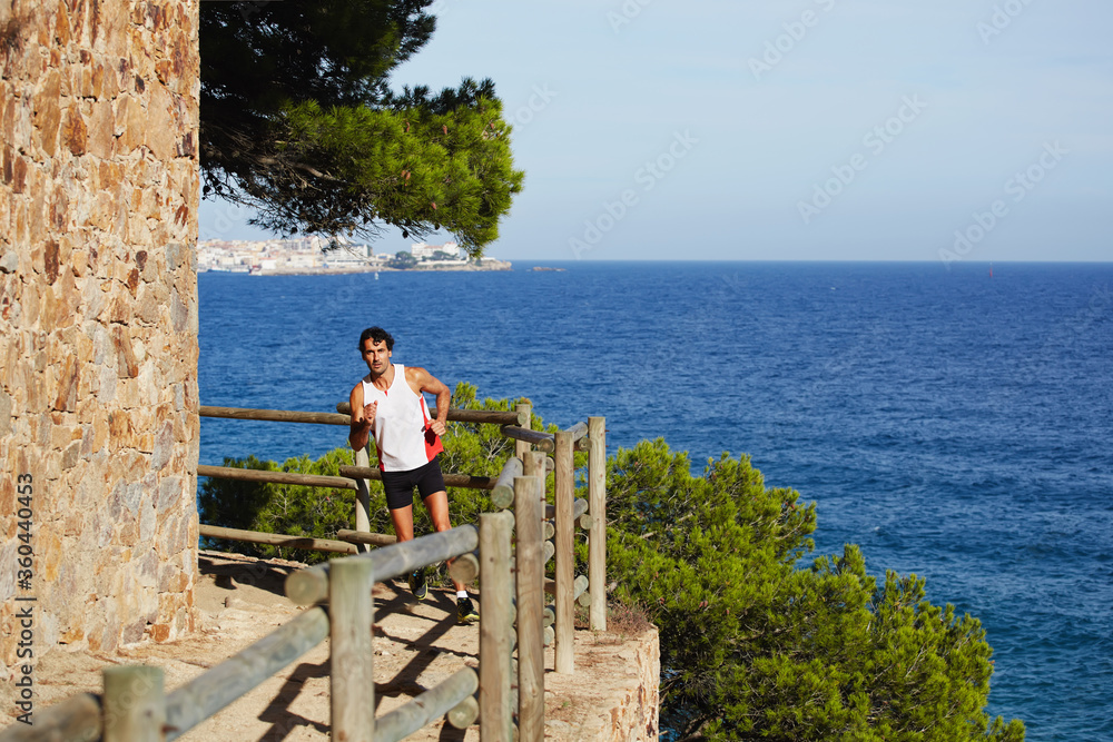 Full length shot of a sporty mature man running on the beach with beautiful sea on background, jogging athlete over mountain trail on high altitude