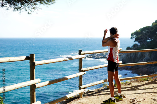 Side shot of handsome young runner stretching his arms before starting his run while standing on edge of a cliff with a wooden fence and enjoying ocean view from altitude © BullRun