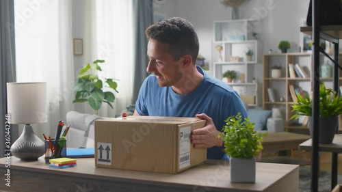 A handsome young man enters the living room with a corton box, sits at the desk and begins to open the parcel