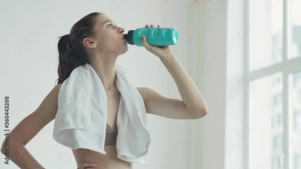 Medium shot of slim Caucasian sportswoman standing in gym with white towel around her neck and drinking water from sport bottle Medium shot of slim Caucasian sportswoman standing in gym with white towel around her neck and drinking water from sport bottle