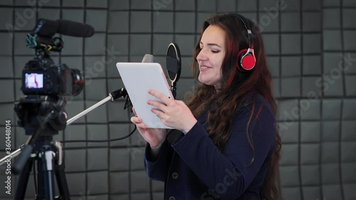 Medium shot of a female voice artist doing voice over in a sound studio. Voice actress reads text from digital tablet and dubbing to the microphone. Podcast production on the sound recording studio