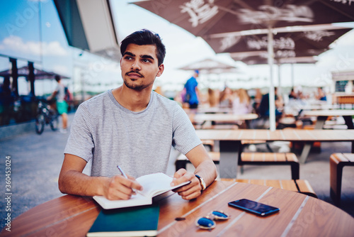 Handsome male designer pondering idea for project making notes and plannings sitting outdoors on terrace of cafe, pensieve hipster guy creating checklist of targets in dairy making schedule on break photo