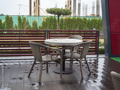 A table and chairs stand on the wet wooden floor of a street cafe against the background of trees and a building under construction
