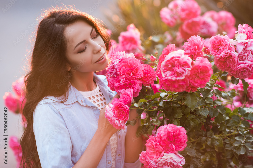 Fototapeta premium Smiling beautiful woman 20-24 year old smelling pink roses in garden outdoors closeup. Summer season. Romance.