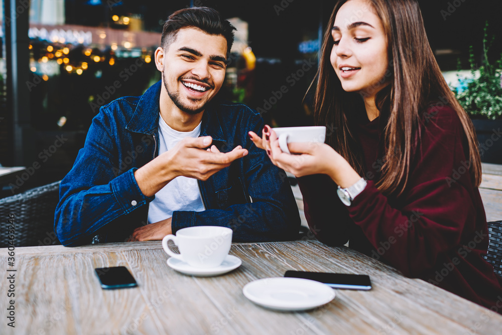 Cheerful male and female teen best friends sitting on cafe terrace drinking coffee and joking, romantic couple in love dressed in casual wear having fun during date communicating and resting