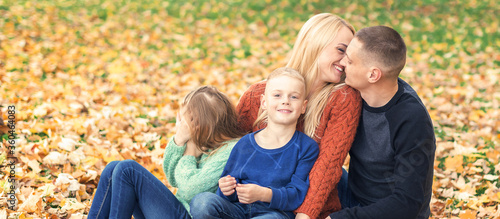 Portrait of young family sitting in autumn leaves. Parents kissing and sitting with children in the autumn park