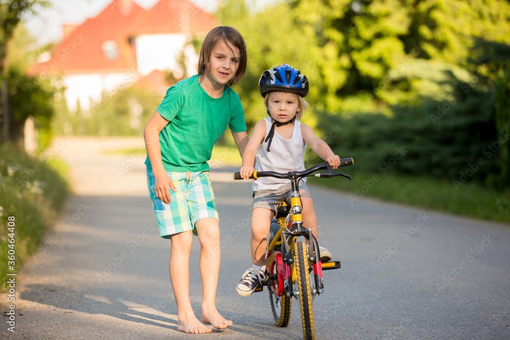 Naklejka premium Older brother, helping his little brother to learn how to ride a bike, holding him and teaching him