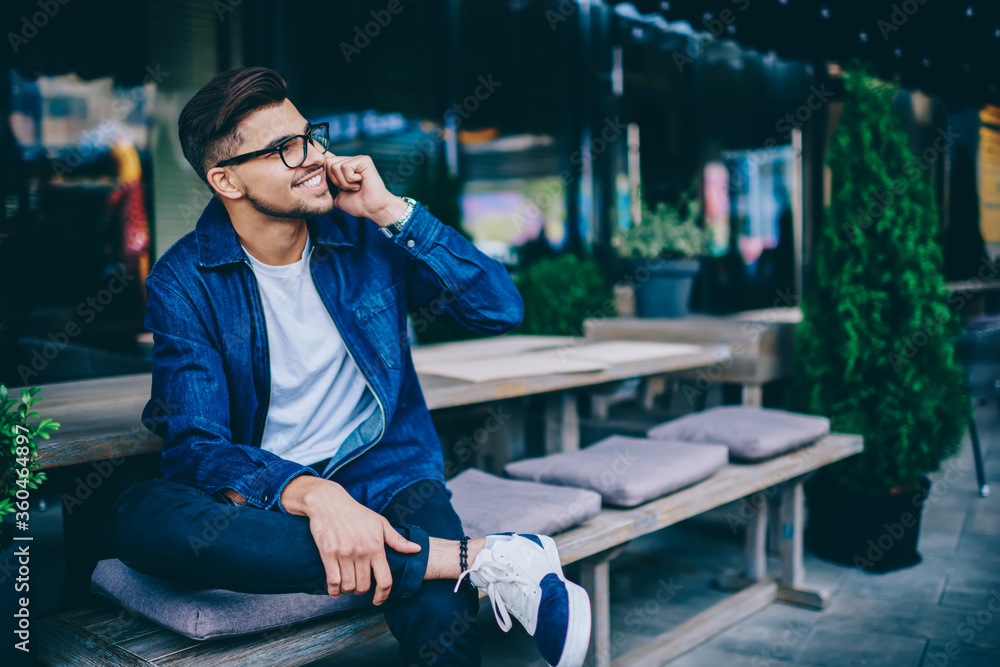 Fototapeta premium Cheerful caucasian male in spectacles having mobile phone conversation sitting outdoors, happy hipster guy in trendy wear satisfied with connection in roaming making telephone call and smiling