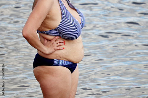 Overweight woman in swimsuit with fat belly standing in a water. Vacation on sea beach, overeating and weight loss concept photo