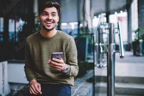 Happy bearded hipster guy excited with good news receiving message on smartphone sitting on urban settings, cheerful handsome man satisfies with mobile tariffs for calls and messages chatting on phone photo