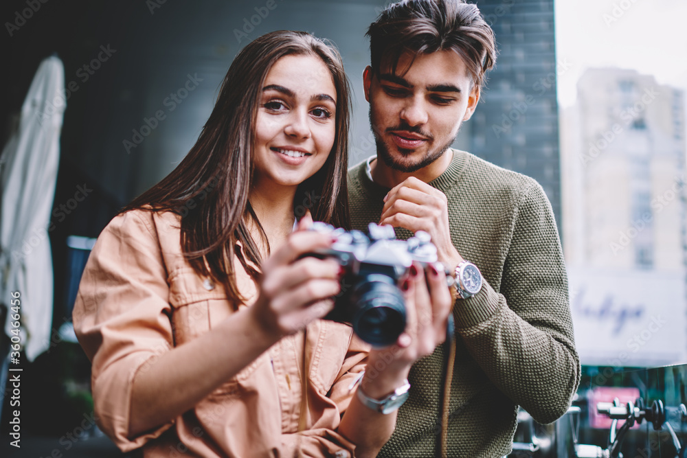 Portrait of cute hipster girl holding vintage camera while her boyfriend making settings for making photos, female amateur asking male photographer friend for solving problems with equipment