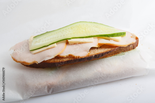 isolated close up shot of a turkey ham and cheese sandwich on white bread with a slice of cucumber on top, lying on a baking paper wrap package on a white background