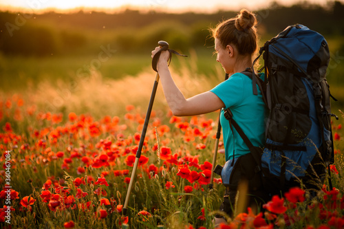 Young woman with backpack and walking sticks stands on field of poppies