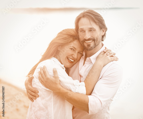 Beautiful couple stand embracing against sea or ocean background. Smiling man and woman in white clothes hugging each other. Tinted image.