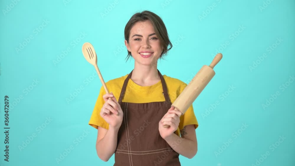 A beautiful young woman wearing an apron is showing cutlery isolated over blue background A beautiful young woman wearing an apron is showing cutlery isolated over blue background