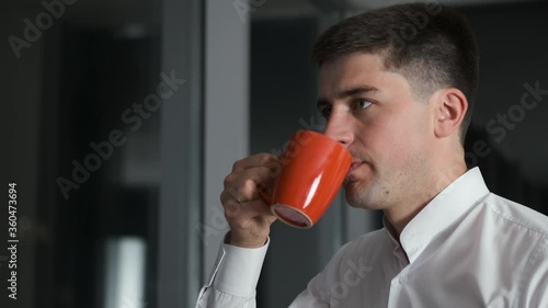 Handsome caucasian successful businessman man lingering at work and standing in evening office near panoramic window overlooking night city and drinking coffee or tea