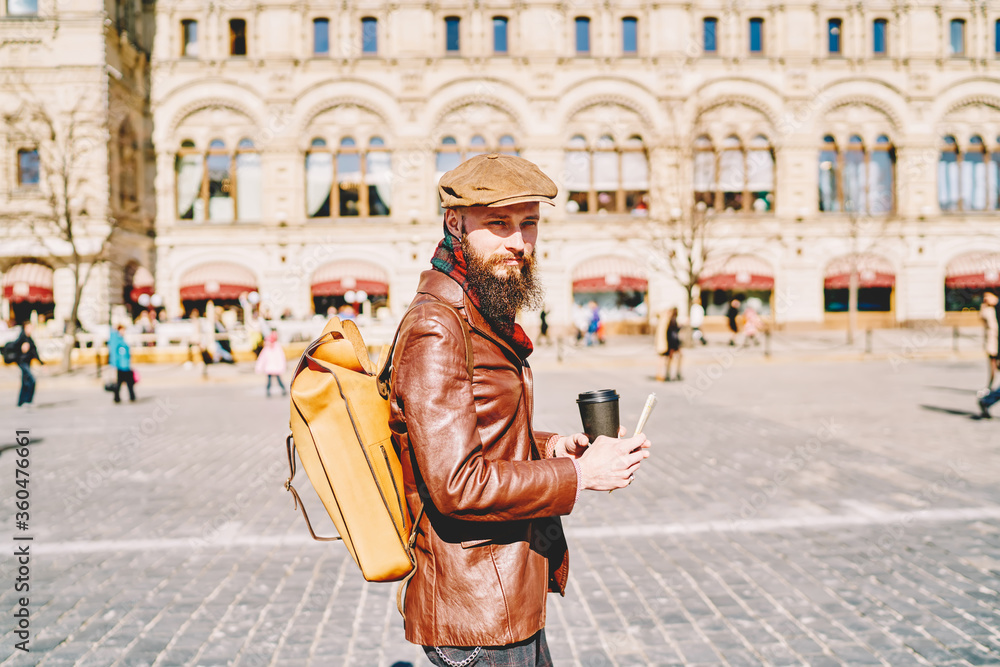 Handsome man in fashionable leather jacket walking on vacation and enjoying sunny autumn day, young bearded male traveller with backpack and takeaway coffee spending holidays on Moscow streets