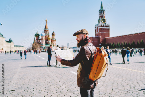 Caucasian male traveller with trendy backpack walking over old Moscow city holding paper map for learn new showplaces, young man dressed in stylish apparel choosing route for trip during sunny day