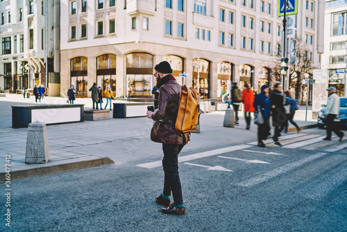 Back view of male tourist with trendy backpack for travelling crossing city road and walking to destination in Moscow, hipster guy in leather jacket strolling around modern streets in Russia