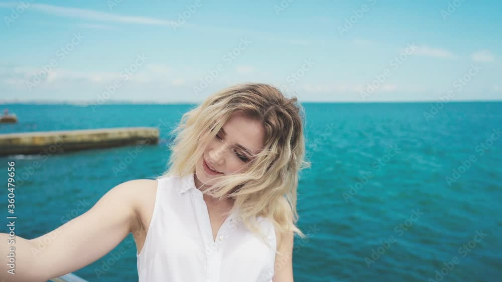 Young attractive blonde girl in a white blouse and blue jeans, makes selfie standing on the pier on sunny summer day Young attractive blonde girl in a white blouse and blue jeans, makes selfie standing on the pier on sunny summer day
